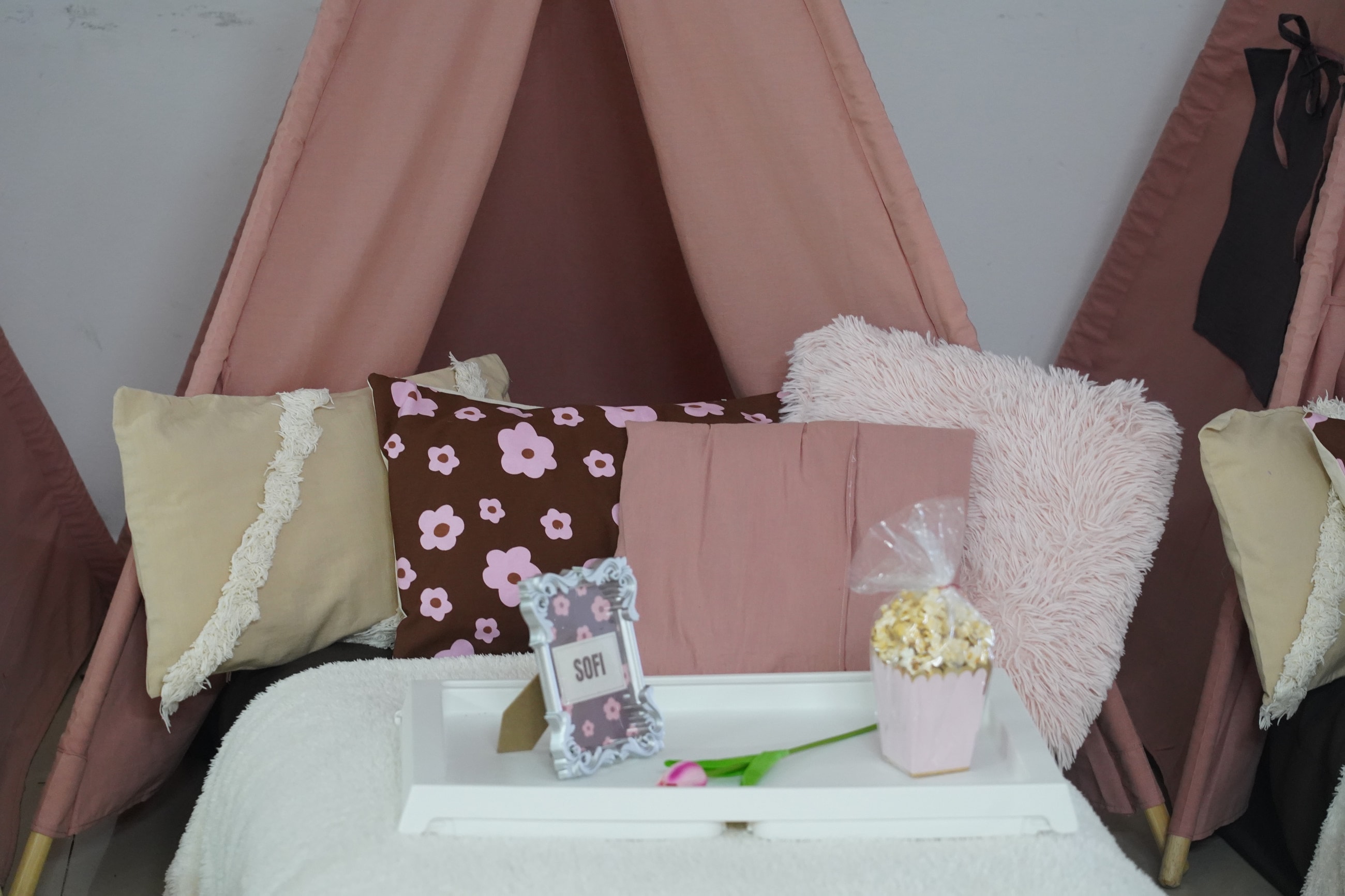 Bandeja blanca con portarretrato, tulipán y pochoclos sobre un colchón, frente a una carpa rosa con almohadones floreados y mullidos.