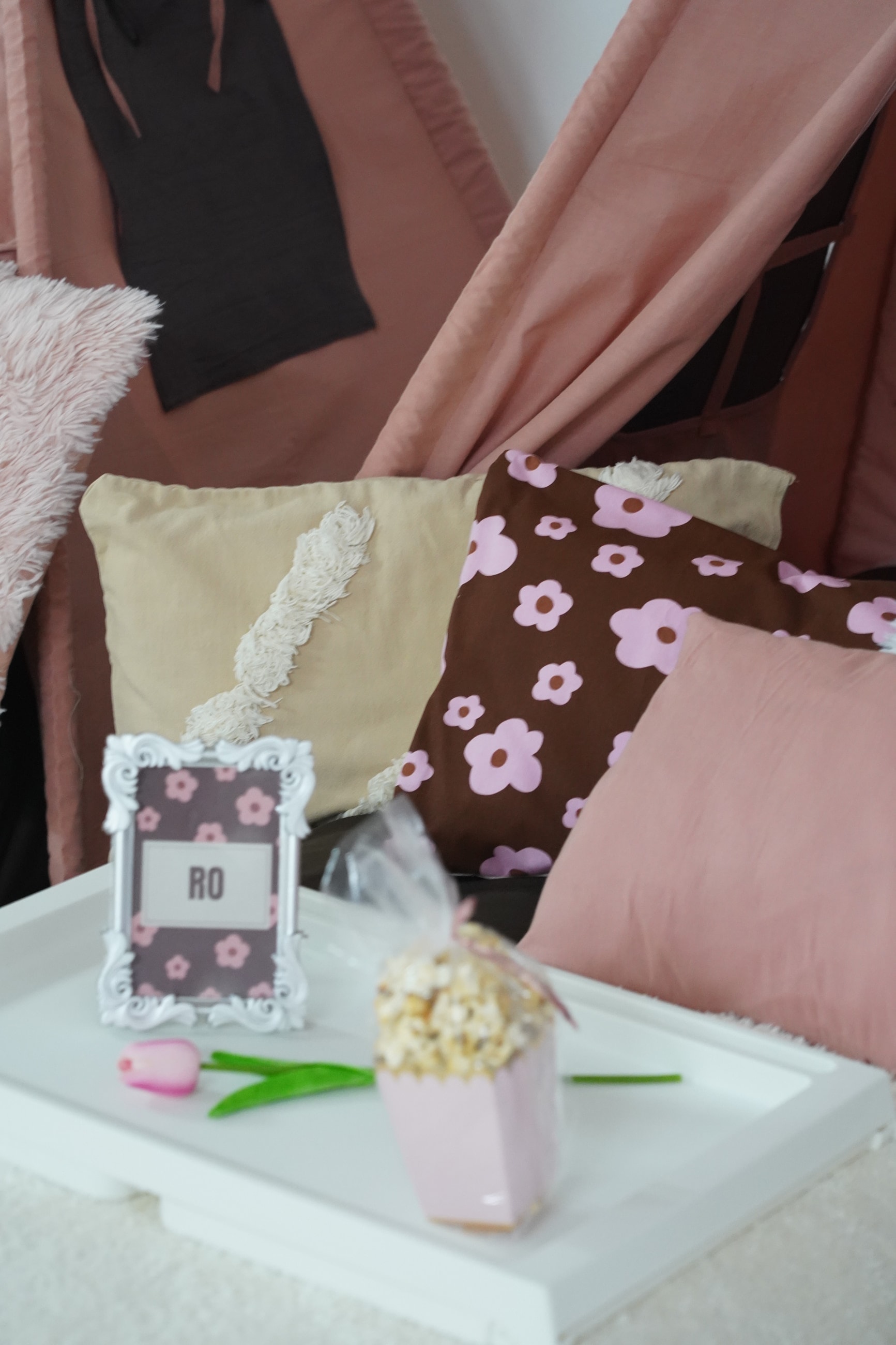 Bandeja individual con portarretrato, tulipán y pochoclos, delante de almohadones beige, floreados y rosa.