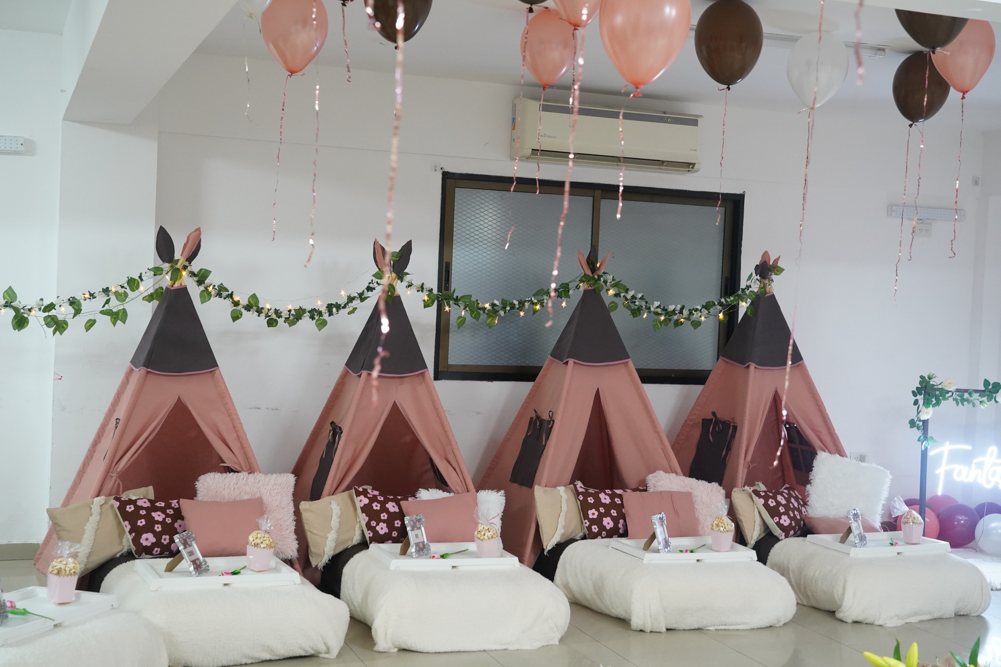 Cuatro carpas tipi rosa y chocolate en fila, con guirnalda de hojas y luces, colchones blancos, almohadones floreados y bandejas individuales.