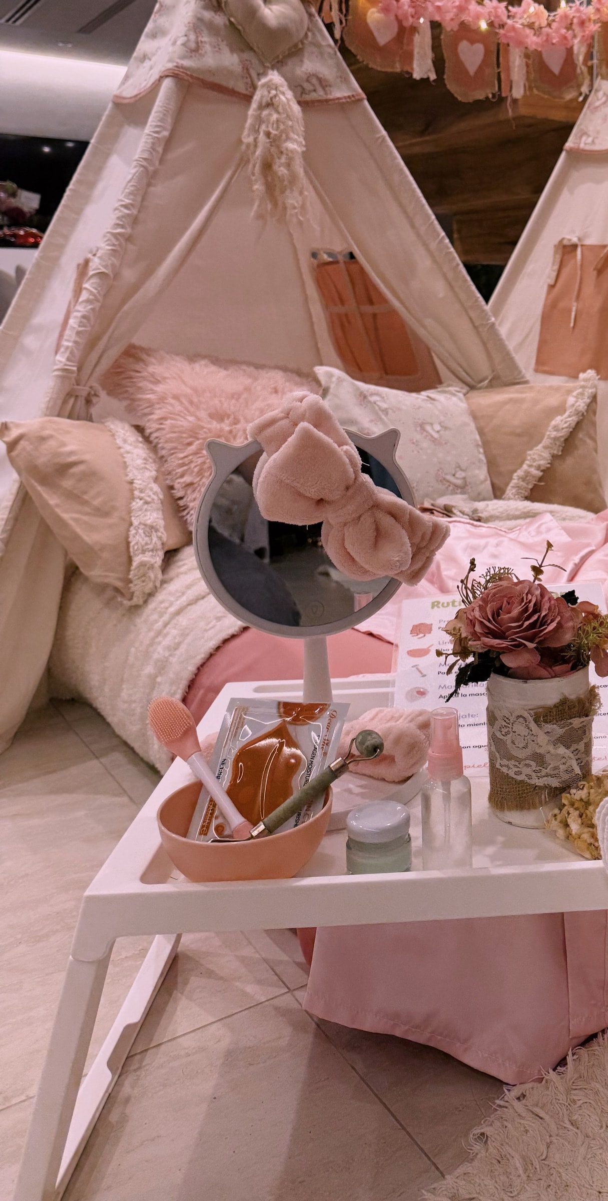 Bandeja del kit de spa con espejo con luces, vincha de moño, bowl, cremas y la rutina de skin care, frente a carpas rosas.